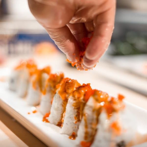 A gloved hand sprinkles bright orange roe over a row of sushi rolls on a white plate.
