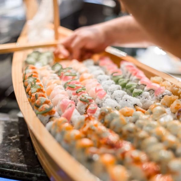 A chef's hands arranging a large variety of colorful sushi rolls on a wooden boat platter.
