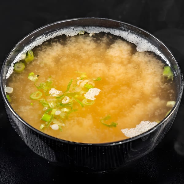 A bowl of miso soup with chopped green onions and white foam on a dark background.
