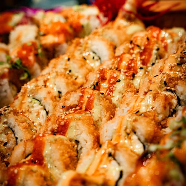 Rows of sushi rolls with tempura flakes, sesame seeds, and drizzled sauces.